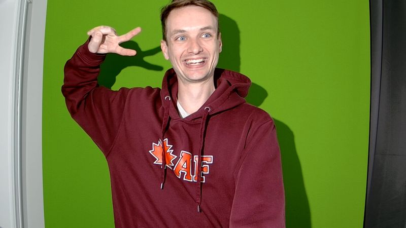Cody Johnston grinning and flashing a peace sign in a maple leaf hoodie in front of a green screen